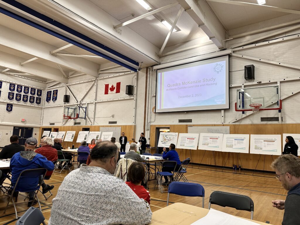 A person presenting in front of others at a gymnasium with power point on screen and presentation panels.