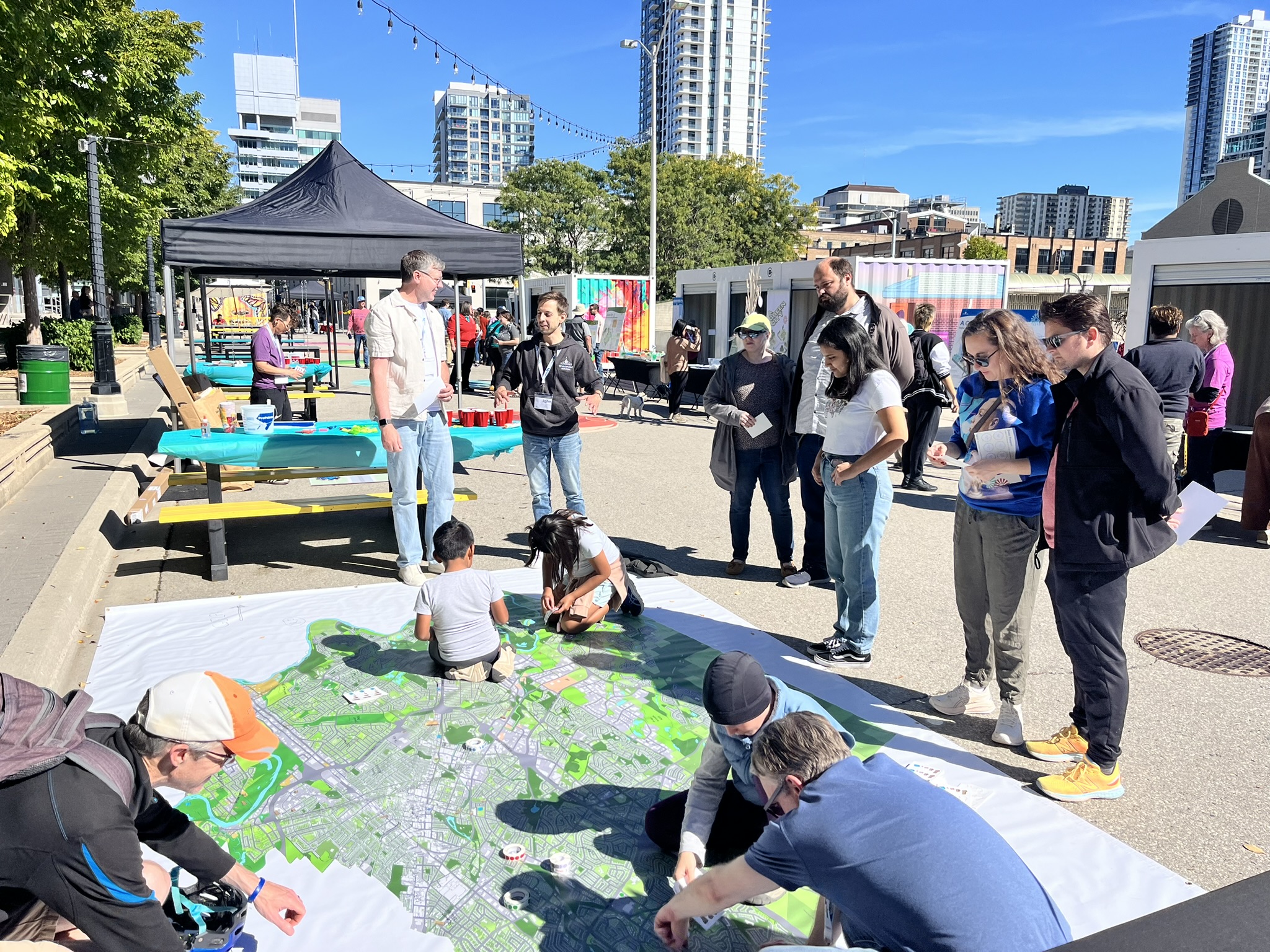 People kneeling and placing stickers on the How We Grow map at the Block Party engagement event, while other people stand around the map watching and discussing.